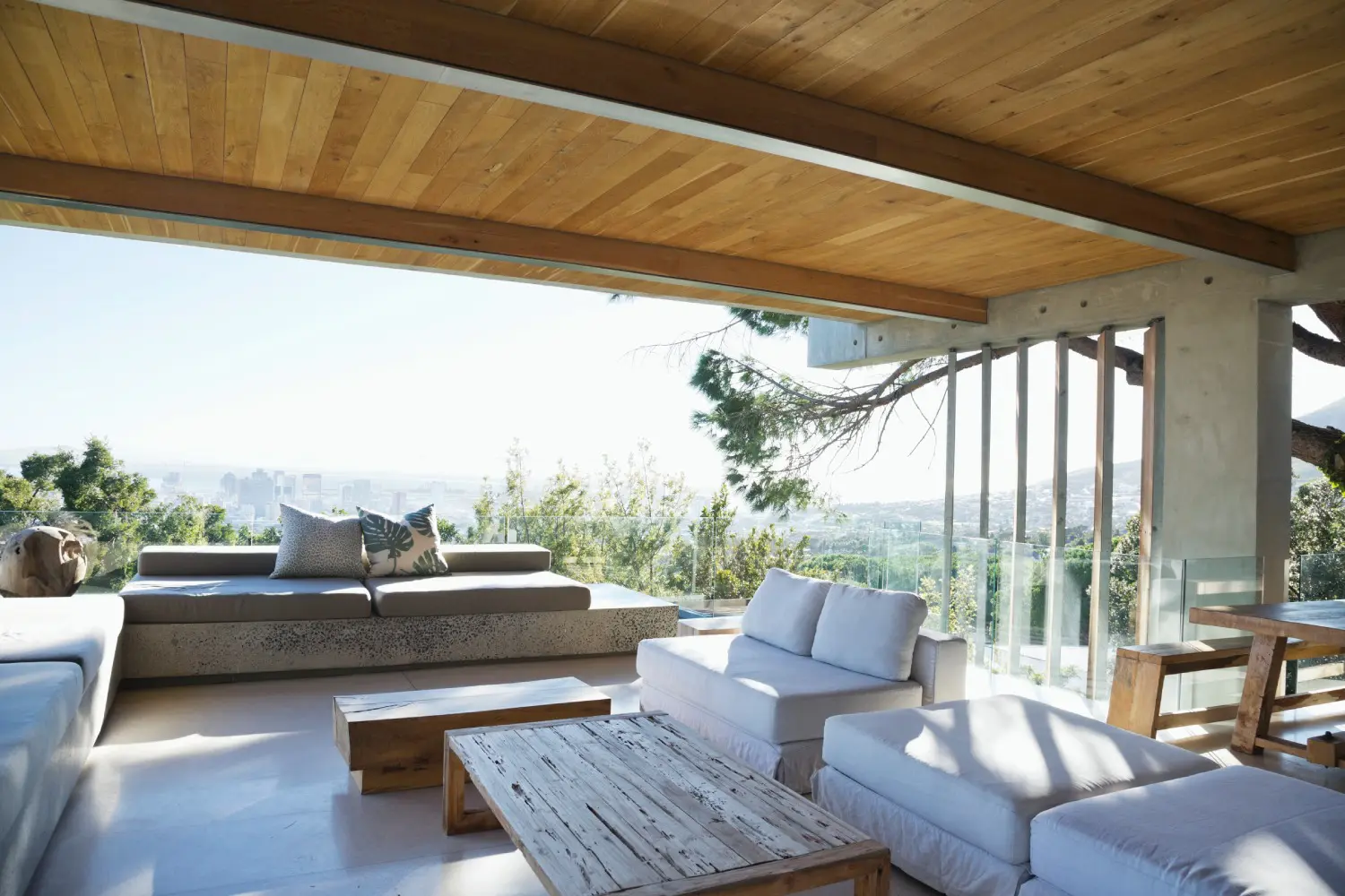 Open-air living room with wooden ceiling and rustic furniture, featuring a view of greenery and a distant cityscape through large windows.