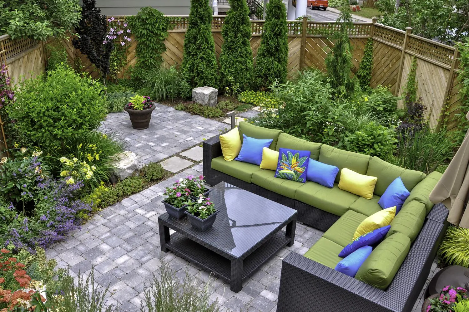 A backyard with a patio features a green and blue sectional sofa set, a black table, potted plants, and lush greenery with wooden fencing.