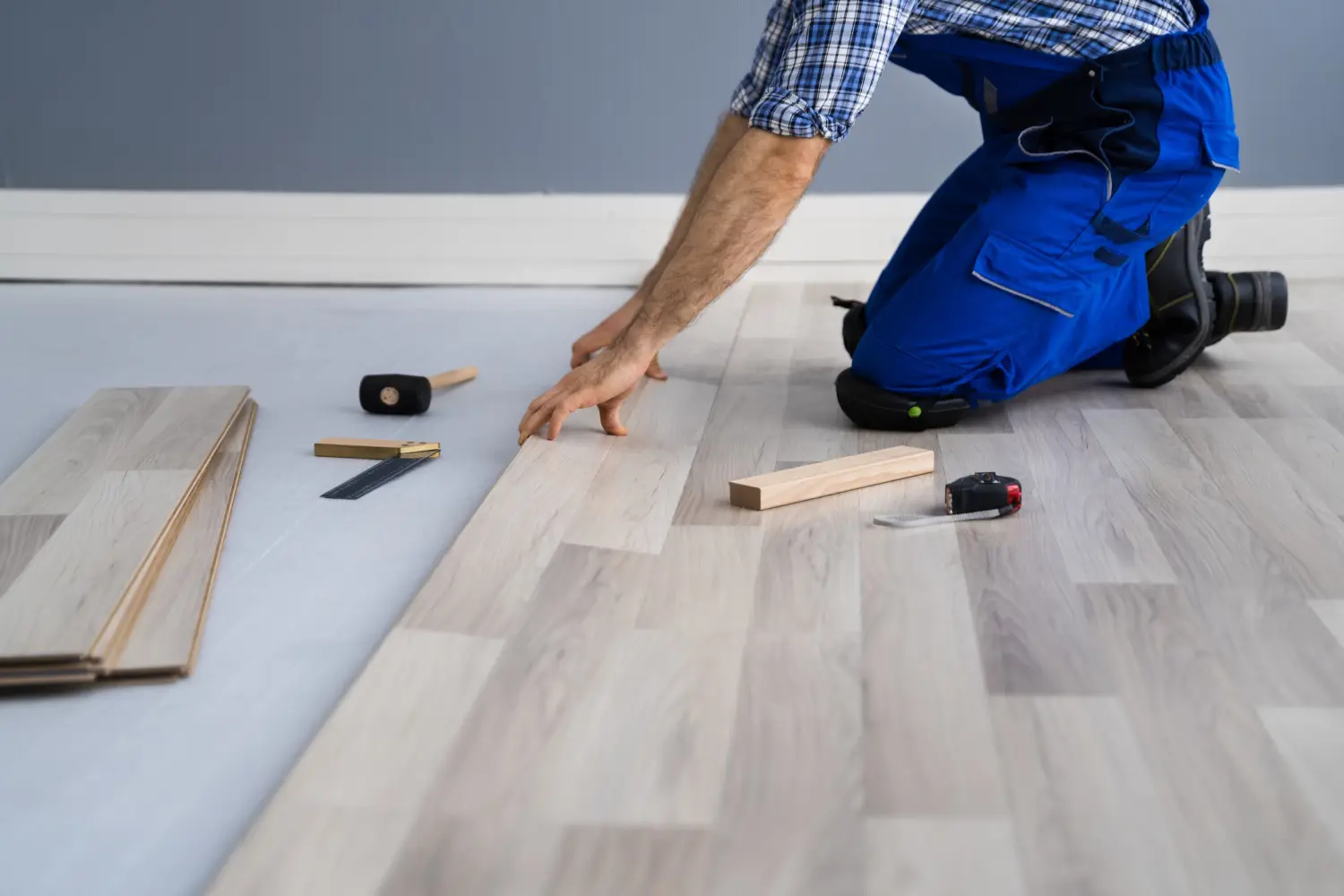 Person kneeling on the floor installing light-colored laminate flooring with tools and wooden spacers nearby.