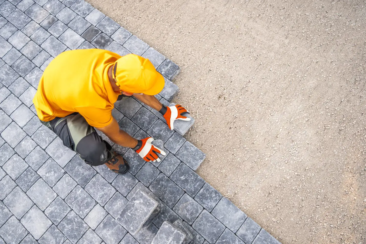 Worker in an orange shirt and gloves laying gray paving stones on a dirt surface.