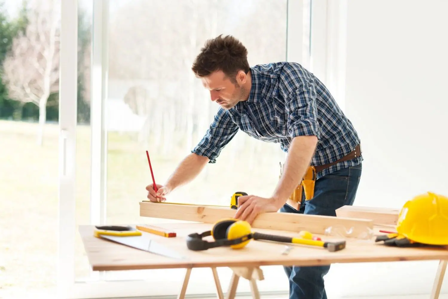 A man in a plaid shirt measures and marks a wooden plank on a table cluttered with tools. Natural light streams through large windows in the background.