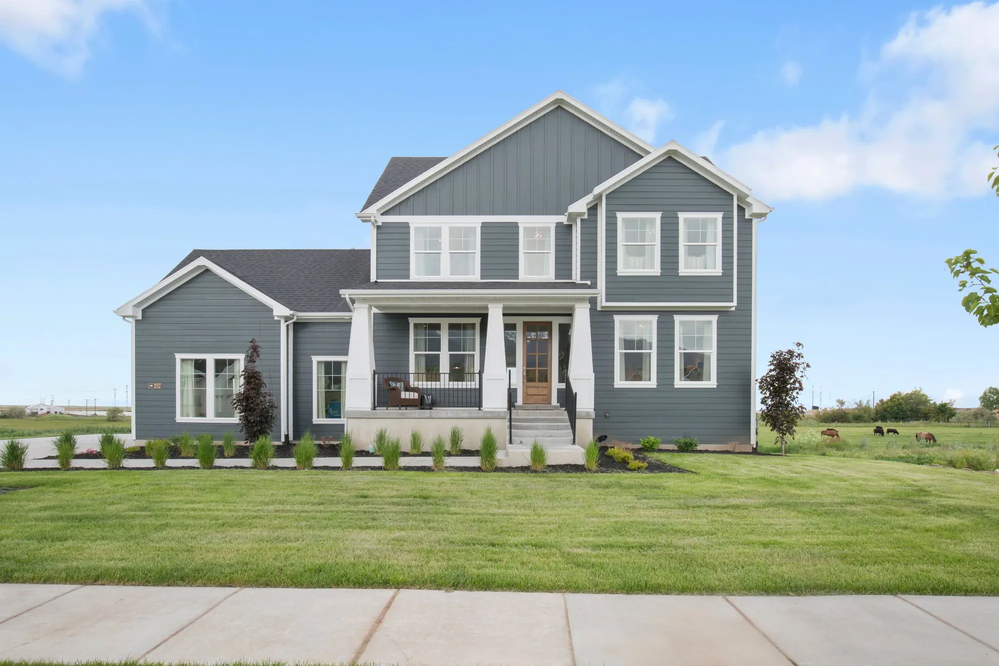 Two-story gray house with a front porch, large windows, surrounded by a green lawn, under a blue sky.