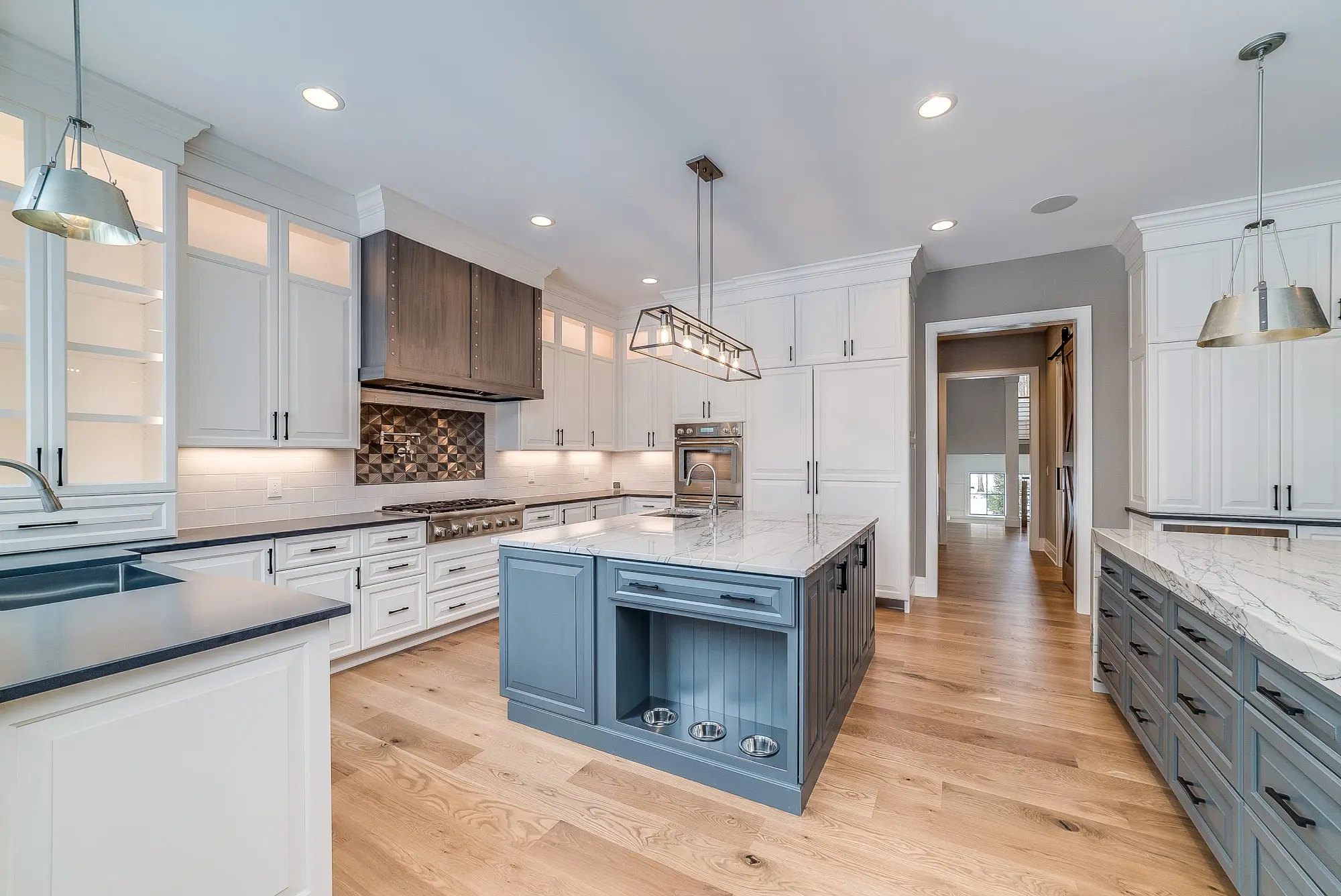 Spacious modern kitchen with white cabinets, light wood flooring, a blue island, stainless steel appliances, pendant lighting, and marble countertops.