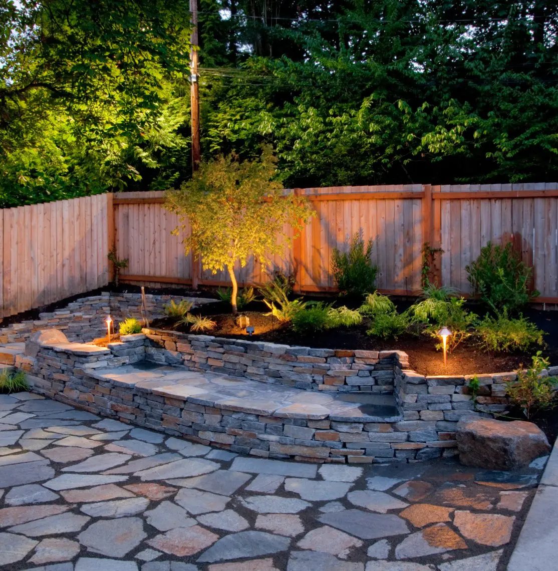 A stone patio with flagstones leads to a raised garden bed. A wooden fence and greenery are in the background, with small lights illuminating the plants and tree.