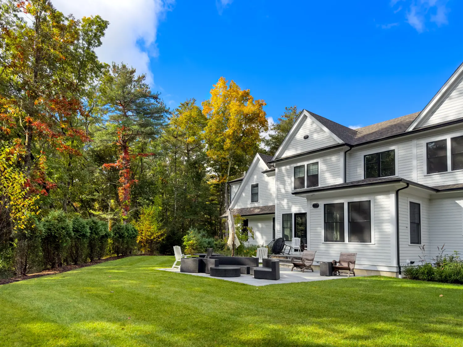 Modern white house with large windows and a black-trimmed roof, surrounded by trees. Backyard features a patio with seating and a grassy lawn under a blue sky.