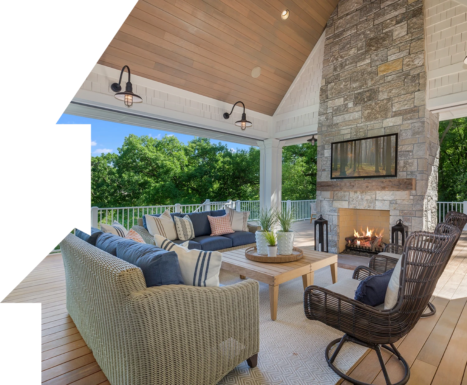 Outdoor patio with wicker furniture, a stone fireplace, and a view of lush greenery. The space has a wooden ceiling, decorative lighting, and a coffee table with plants.