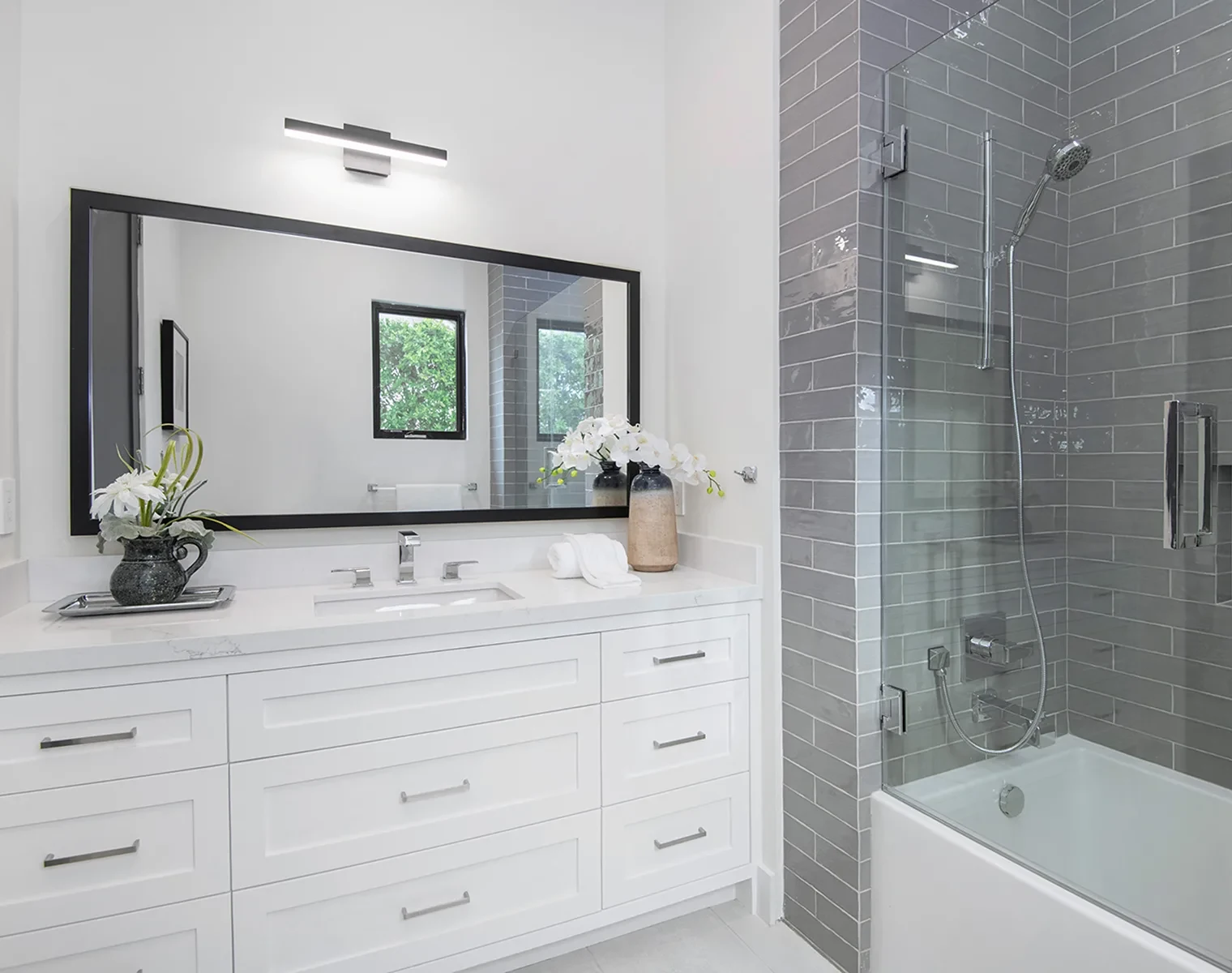 Modern bathroom with white vanity, large mirror, and gray-tiled shower. Two vases with white flowers sit on the counter. A window is reflected in the mirror.